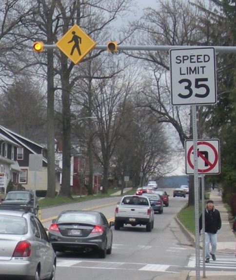 Photo of many cars driving along a busy street where there is featured a designated crosswalk with overhead warning sign and yellow flashing lights.